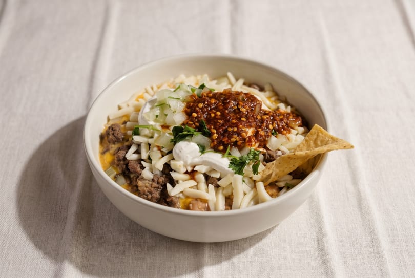 A bowl of chili with shredded cheese, sour cream, onions, and a tortilla chip.